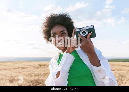 Smiling young woman holding camera in field Stock Photo