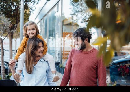 Smiling woman carrying daughter on shoulder by man on sidewalk Stock Photo