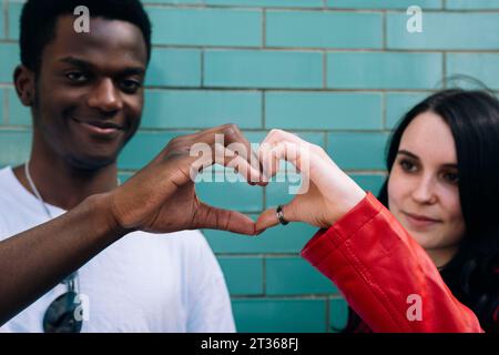 Smiling young couple gesturing heart shape in front of brick wall Stock Photo