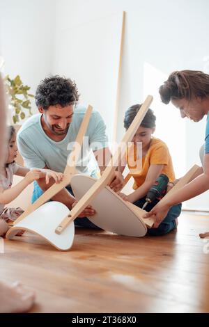 Happy family with woman using drill machine on wooden chair at home Stock Photo