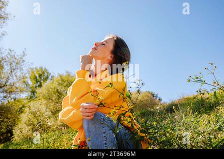 Smiling woman with hand on chin sitting in nature under sky Stock Photo