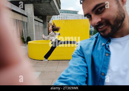 Smiling man taking selfie with woman jumping in background Stock Photo