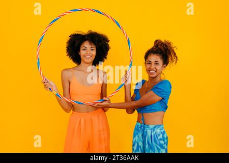 Smiling women holding plastic hoop standing against yellow background Stock Photo