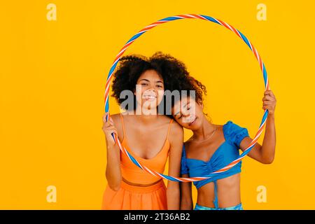 Woman leaning on friend holding plastic hoop against yellow background Stock Photo