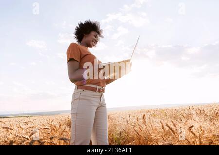 Smiling Afro farmer with laptop standing in barley field Stock Photo