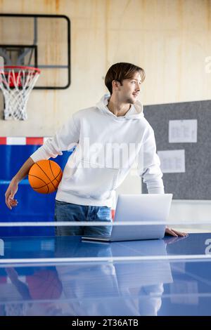 Young trainee standing with basketball and laptop in office Stock Photo