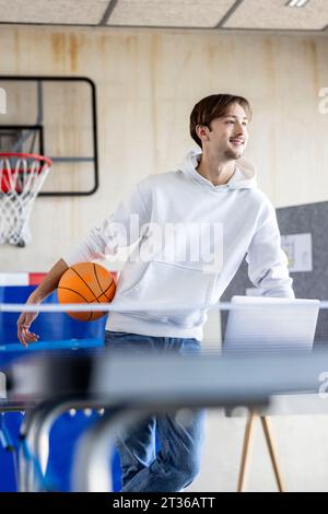 Happy young trainee standing with basketball and laptop in office Stock Photo