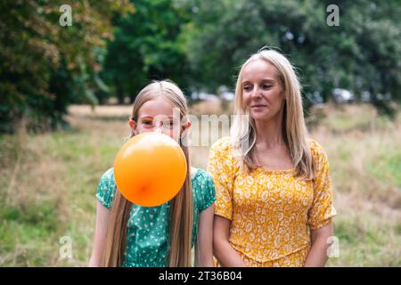 Smiling mother looking at daughter blowing balloon in park Stock Photo