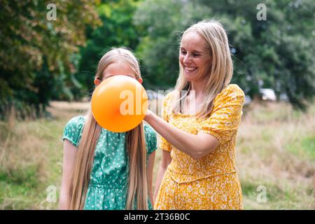 Happy woman helping daughter blowing balloon in park Stock Photo