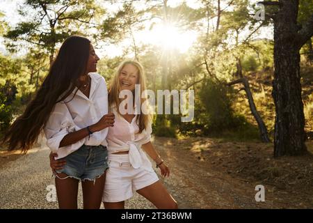 Cheerful mother with arm around daughter walking on road in forest Stock Photo