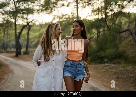 Smiling mother and daughter with arms around walking on road in forest Stock Photo