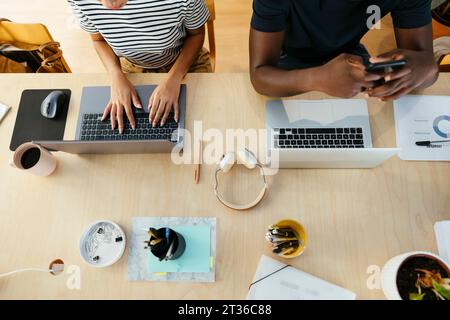 Young student typing on laptop with friend using smart phone at desk Stock Photo