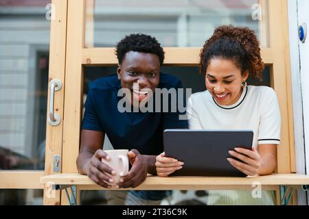 Smiling young student with friend using tablet PC and leaning out of window Stock Photo