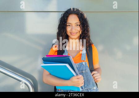 Smiling student holding spiral notebooks in front of wall at campus Stock Photo