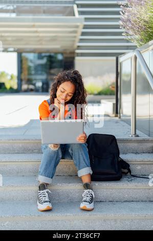 Smiling student with backpack using laptop on steps in campus Stock Photo