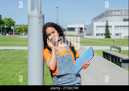 Young student talking on smart phone and holding notebook near pole in campus Stock Photo