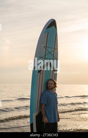 Young man standing in front of paddleboard at beach under sky Stock Photo