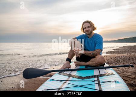 Happy man with tattoo sitting on paddleboard at beach Stock Photo