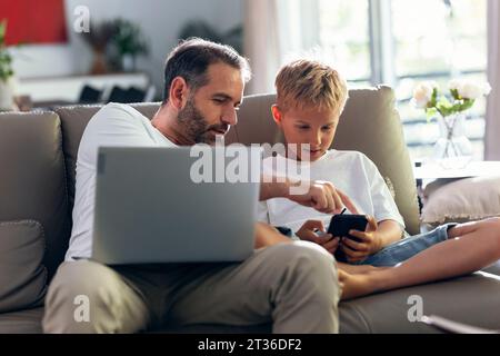 Father and son using mobile phone sitting on sofa at home Stock Photo