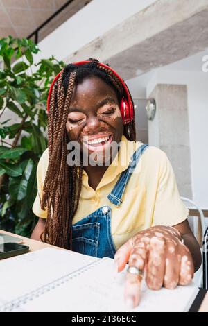 Happy young woman wearing wireless headphones reading book at table Stock Photo