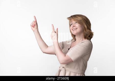 Woman looking up with sly smile and idea pointing at marketing advertisement. Portrait of blonde female in dress, standing on white background Stock Photo
