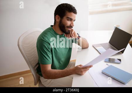 Smiling pensive handsome islamic young man in green t-shirt read documents, work with laptop with empty screen Stock Photo