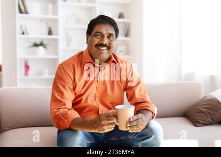 Happy mature indian man sitting on couch, drinking coffee Stock Photo