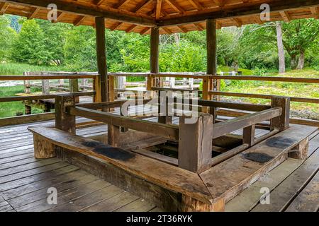 Old gazebo with wooden platform, fences, benches and pavilion-type roof over lake, opening in middle to observe fish, bridges and leafy trees in backg Stock Photo