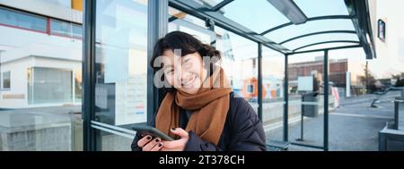 Cute smiling asian girl standing on bus stop, holding smartphone, wearing winter jacket and scarf. Woman commuting to work or university via public Stock Photo