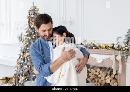 Family couple celebrating new year together on Christmas, standing near Christmas tree in living room smiling joyfully and hugging, man and woman in love. Stock Photo
