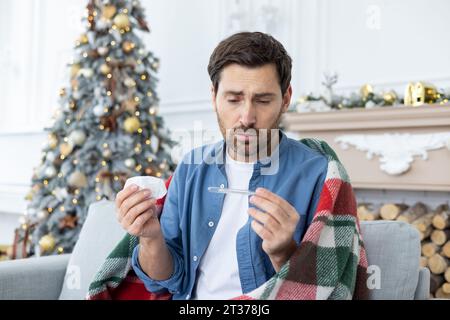 Upset young man sitting at home sick, wrapped in a blanket, holding a napkin in his hand and looking disappointedly at the thermometer. Stock Photo
