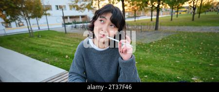 Portrait of asian woman, student in park, sits on bench with digital tablet and a pen, thinking, looking aside thoughtful, making notes, does her Stock Photo