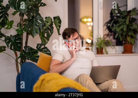Man in protective eyeglasses talks phone works on laptop sits on couch at home in evening. Stock Photo