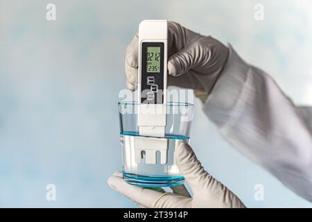 pH meter in hands with gloves, glass of water on a blue background. Measurement of the characteristics of drinking water. The hardness of the water. p Stock Photo