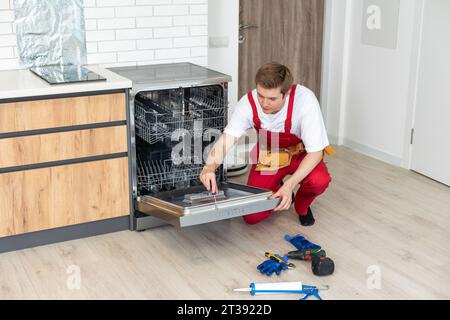 Full length of repairman repairing dishwasher with screwdriver in kitchen. Stock Photo