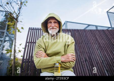 Portrait of elderly man in a hoodie with the hood on his head and arms crossed over his chest, during exercising outdoors. Stock Photo