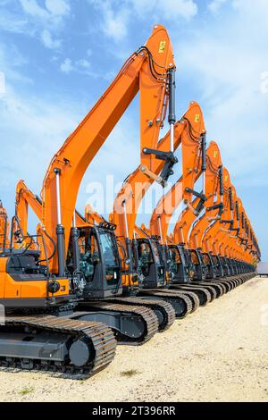 Tens of brand new orange caterpillar excavators lined up in a row outdoors under a stormy sky. Stock Photo