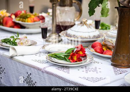 Close-up of a luxurious dining table arrangement showcasing a red flower on a plate, a folded napkin, porcelain dishes with gold detailing, a brass pi Stock Photo