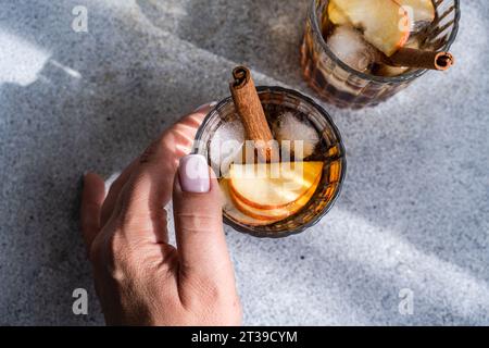 From above of anonymous hand holding a glass of apple cider cocktails adorned with cinnamon stick, star anise, and apple slices, showing fresh ice cub Stock Photo