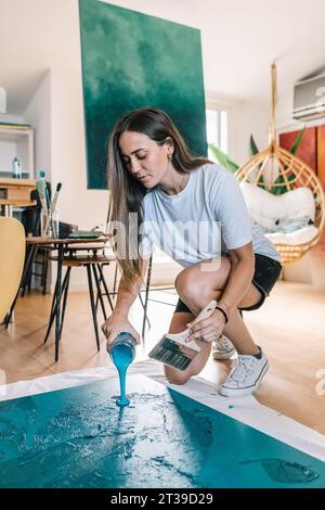 Professional female painter pouring blue liquid paint from plastic container on floorwhile creating artwork during painting at home Stock Photo