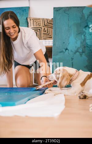 Young smiling female painter in casual clothes painting wooden board while playing with with cute obedient dog holding brush during creation of artwor Stock Photo