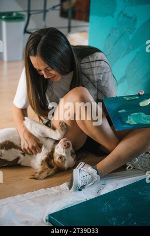 Young happy female artist sitting on floor and playing with adorable dog while holding painting in modern art studio at home Stock Photo