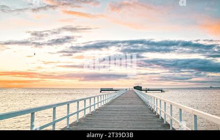 Long wooden pier with metal railings leading to mind blowing rippling seawater view against orange black floating clouds in blue sky during sunset Stock Photo