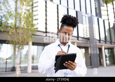 Serious black male entrepreneur in formal white shirt typing with pen on tablet while standing on street with blurred background on sunny day Stock Photo