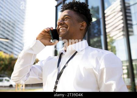 Low angle portrait of cheerful African American male entrepreneur in formal wear speaking on smartphone while standing on street and looking away Stock Photo