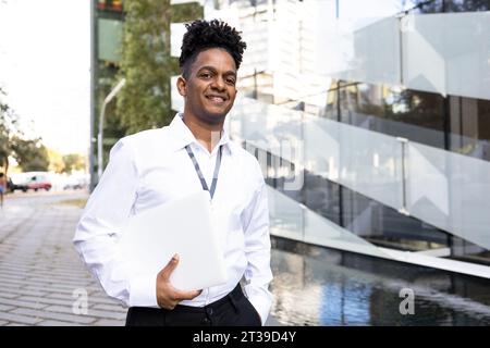 Confident African American male entrepreneur in formal clothes standing near modern building with hand in pocket while holding laptop looking at camer Stock Photo