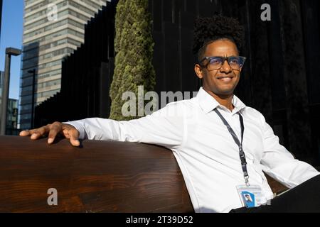 Positive African American male in white shirt and shaved dreadlock sitting on concrete chair with back rest on city street on sunny day Stock Photo
