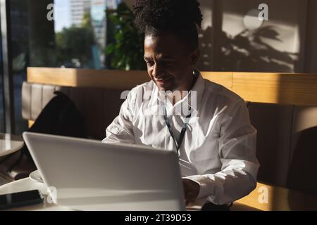 Happy young African American male freelancer in casual clothes and shaved dreadlocks sitting at cafe table while browsing laptop Stock Photo
