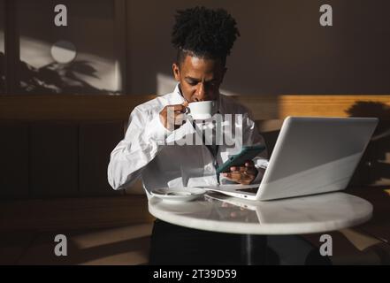 Concentrated African American male freelancer in casual clothes sitting at table with laptop and drinking hot coffee while working on remote project Stock Photo