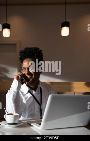Positive African American male freelancer in casual clothes sitting at table with laptop and hot coffee while talking on mobile phone and working Stock Photo